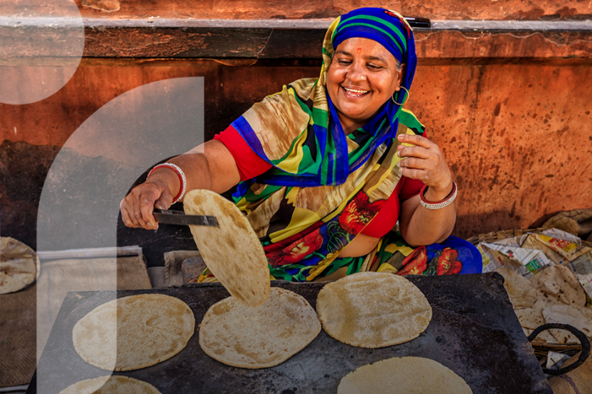 A smiling woman wears colorful clothing while cooking street food on an open air griddle