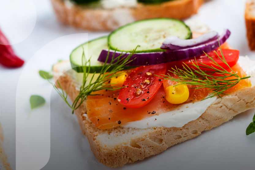 A slide of white bread topped with a white spread, salmon, tomatoes, cucumber, corn, red onion and dill on a white table