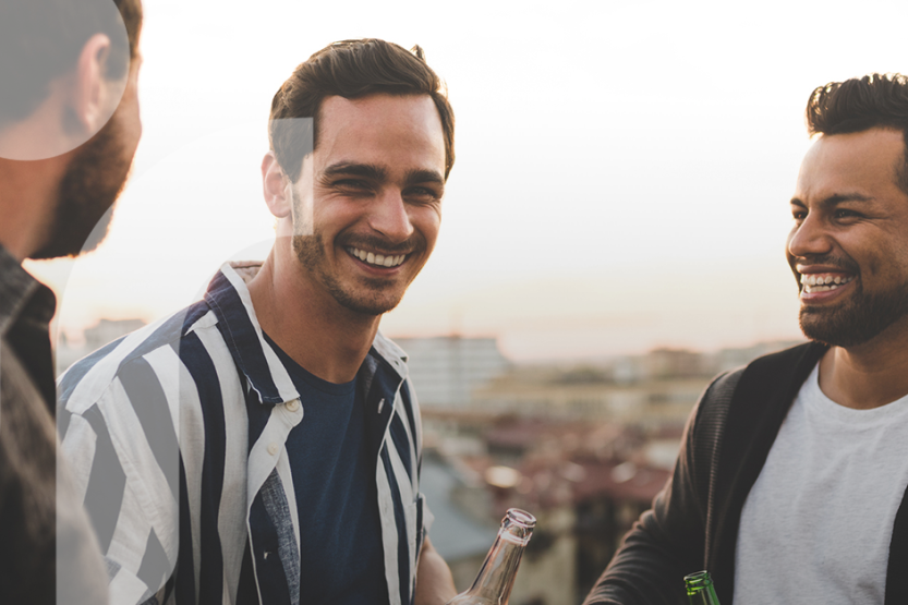 Three men in casual clothing are laughing together with a setting sun in the background 