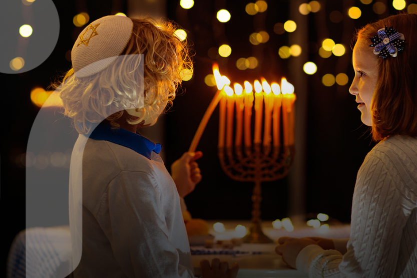 A young boy and girl wearing white with blue accents light a menorah with a night sky background