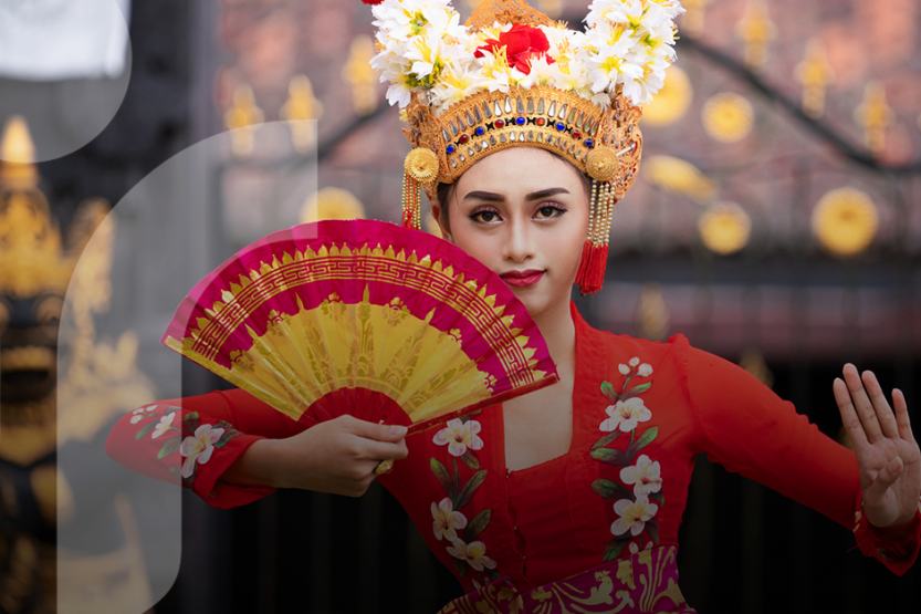 A woman in traditional Indonesian makeup wearing a red dress, gold head piece, and carrying a red and yellow fan, does a traditional dance