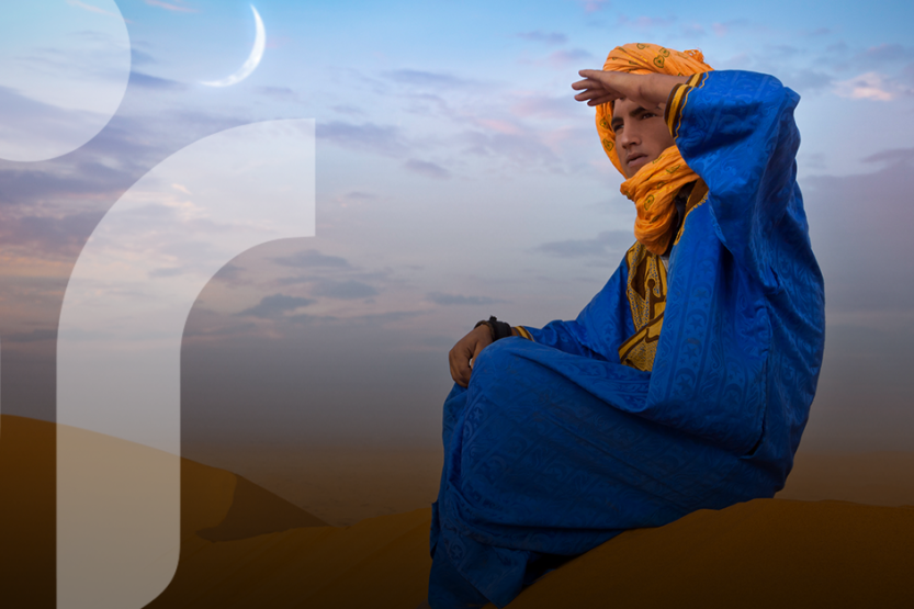 A Berber man in a desert setting sits on a sand dune looking into the horizon wearing a blue tunic and yellow head covering 